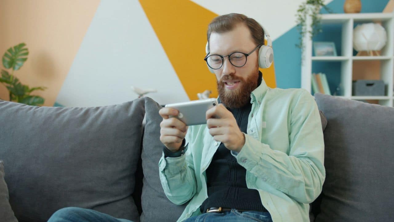 A bearded man with headphones watches content on a smartphone while sitting on a couch.