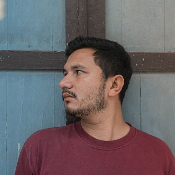 A man in a red shirt stands looking to the side against a blue wooden wall.