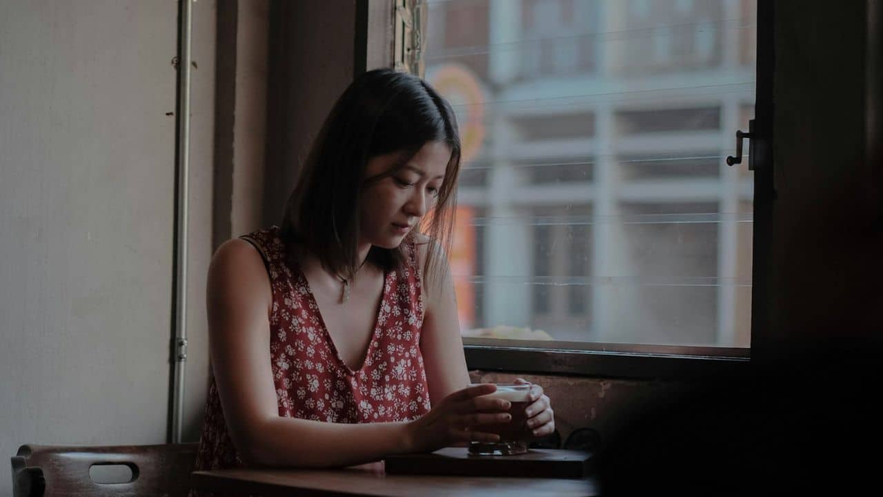 A young woman in a red floral top sits by a window holding a glass.