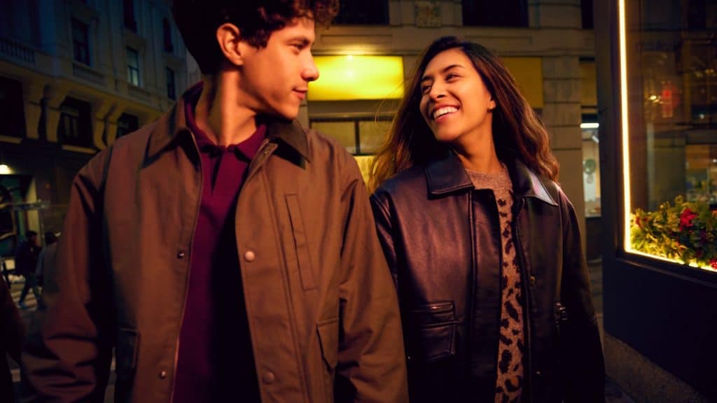 A young man and woman are walking together on a city street at night, smiling and looking at each other.
