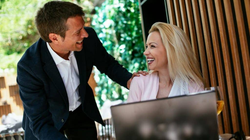 A man smiling while greeting a woman sitting at a laptop.