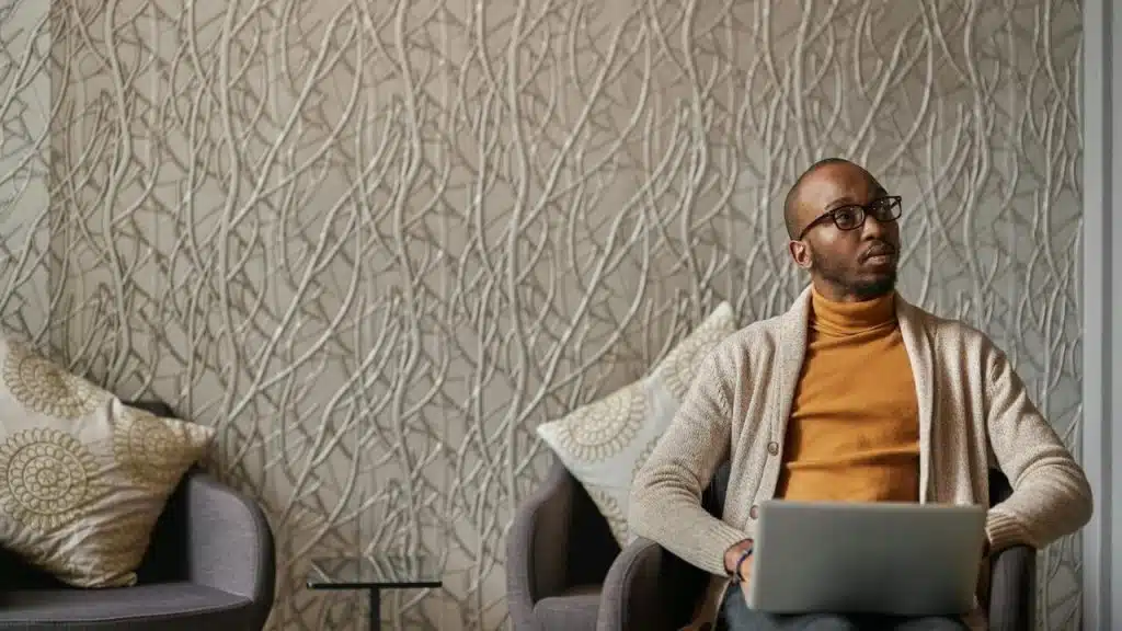 A man sitting with a laptop, looking to the side thoughtfully.