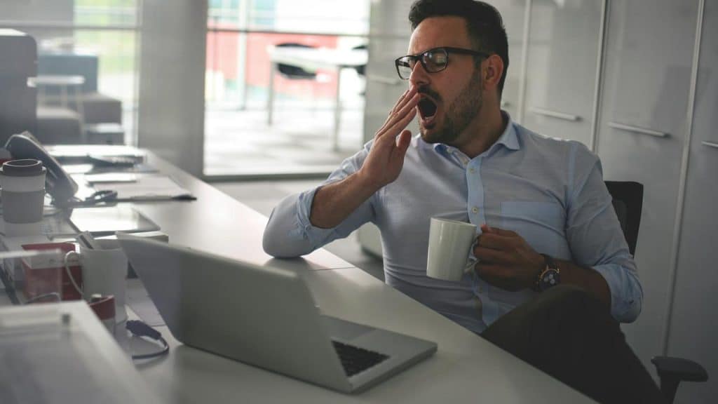 A man in glasses yawning while holding a coffee mug at his desk.