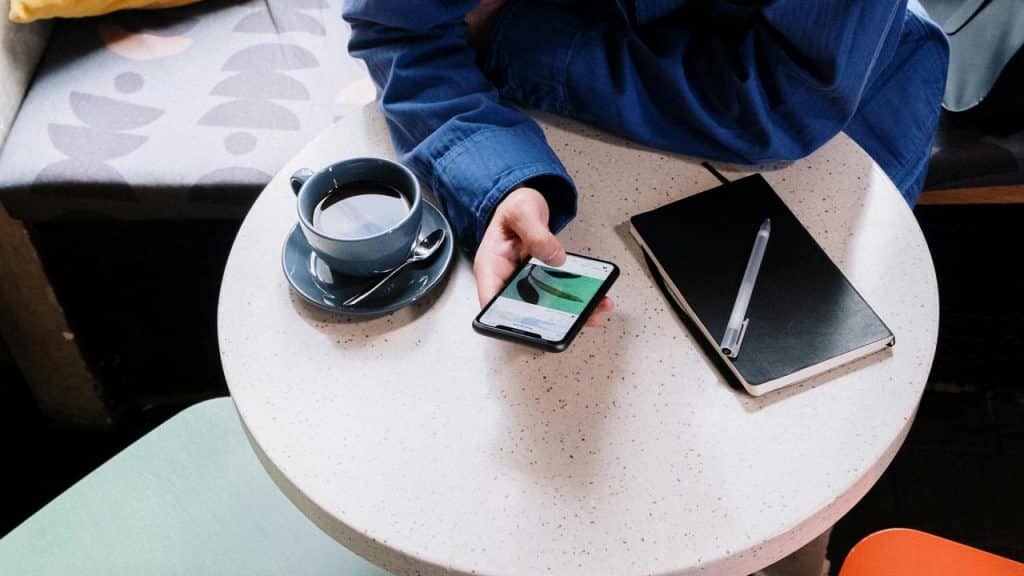 A person sitting at a café table holding a phone beside a cup of coffee and a notebook.
