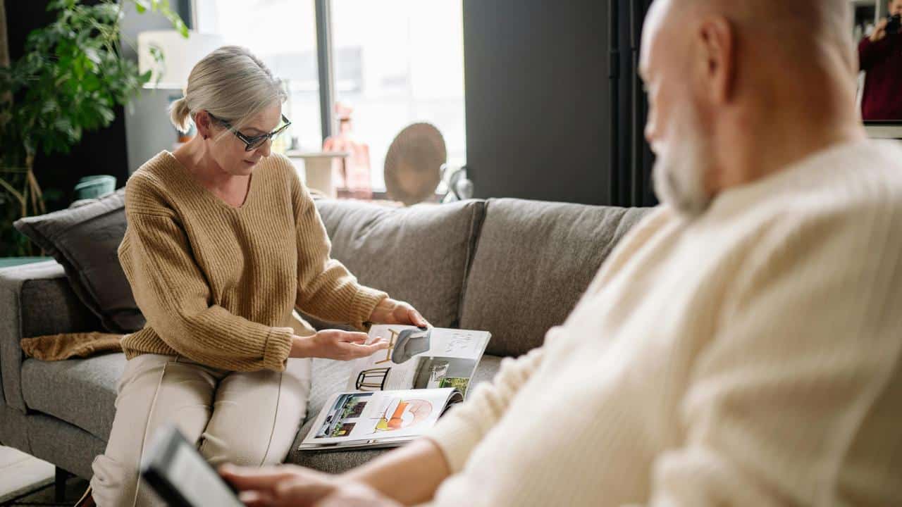 An older woman showing a magazine to a seated man on a couch.