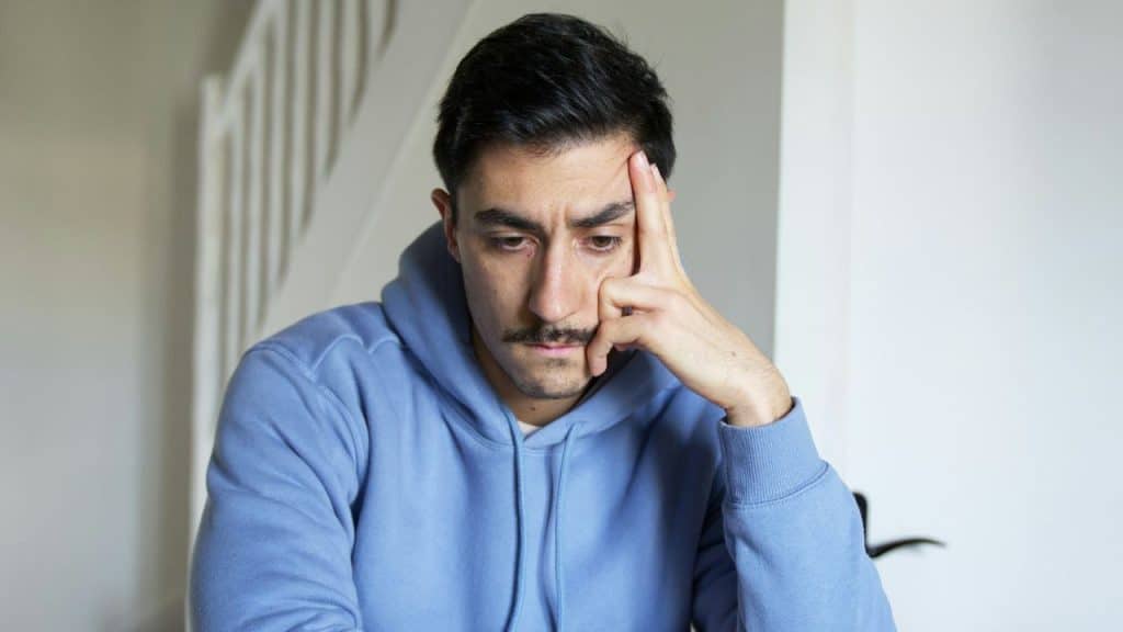 Man in a blue hoodie, resting his hand on his forehead, looking down.