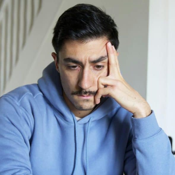 Man in a blue hoodie, resting his hand on his forehead, looking down.