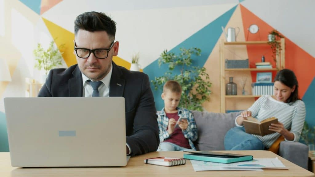 A man works intently on a laptop; a child and woman read on a sofa behind him.
