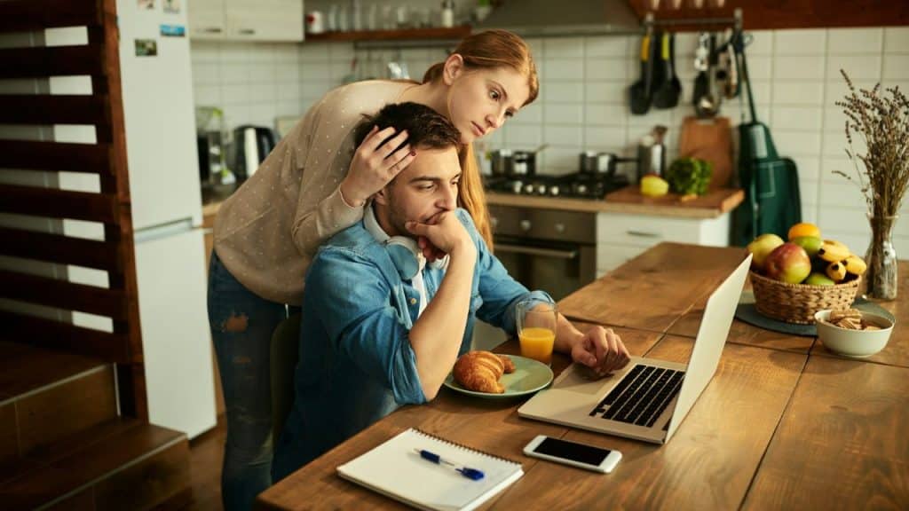 A woman comforts a man working on a laptop at a kitchen table.