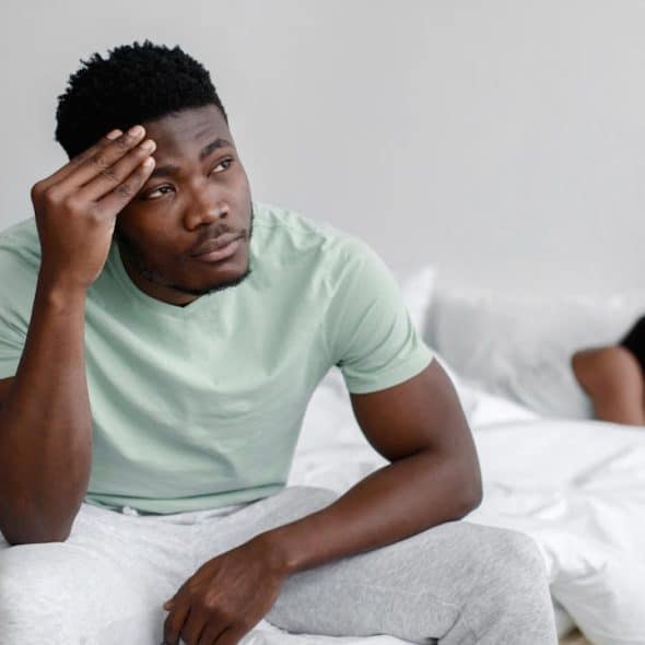 A man sits on a bed looking stressed with his hand on his forehead, while a woman lies facing away from him in the background