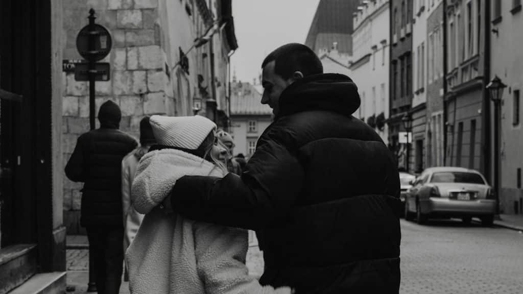 A black and white image of a couple embracing on a cobblestone street in a European city, looking at each other.