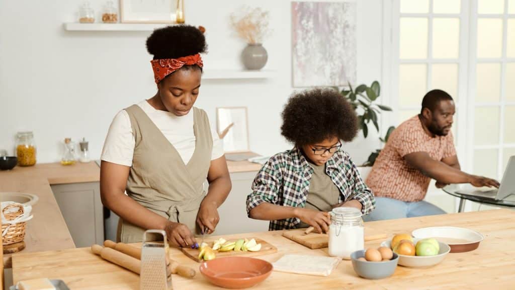 A woman and her son are cooking something in the kitchen while a man works on his laptop in the background.