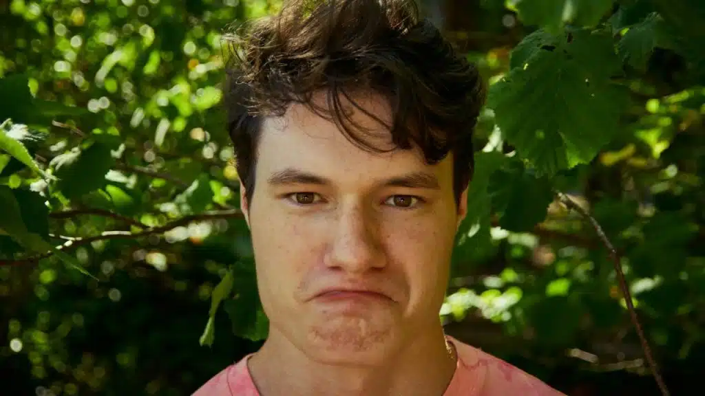 A young man with a slight frown and pouting lips is standing near green foliage.