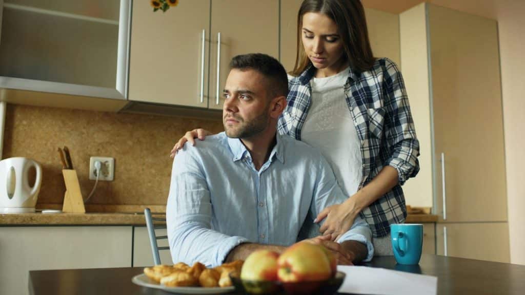 Woman standing behind a pensive man sitting at a kitchen table with food and a mug.
