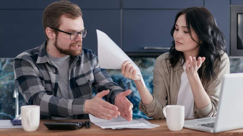 Man and woman arguing over papers at a table with a laptop and coffee cups.