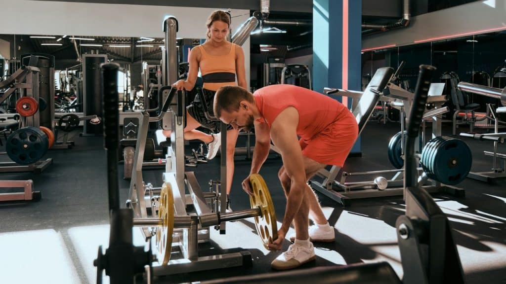 A man loads a yellow weight onto a machine while a woman watches him at the gym.