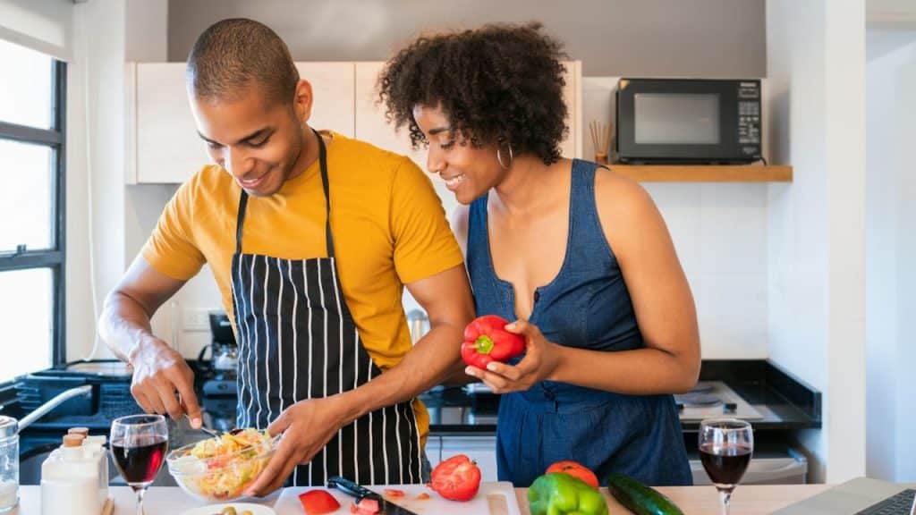 Smiling couple cooking in a bright kitchen, the man stirring a salad while the woman holds a red bell pepper.