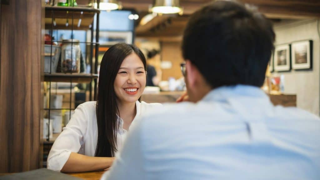 Smiling young woman with long dark hair talking to a man across a table in a cafe or restaurant.