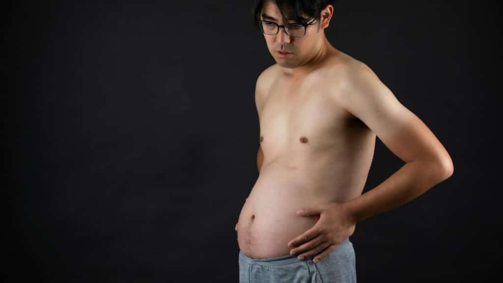 A shirtless man in glasses looks down at his stomach, holding his side against a dark background.