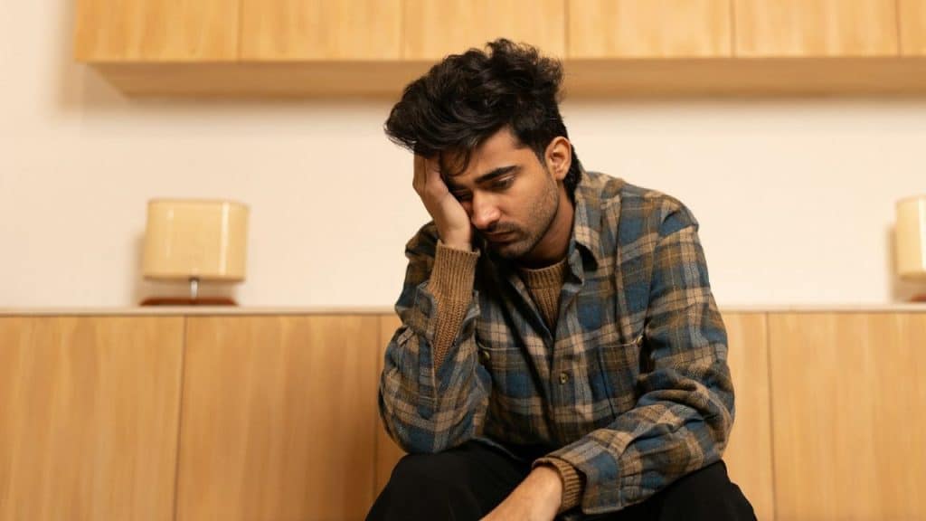 Young man in a plaid shirt with his head in his hand looking down sadly.