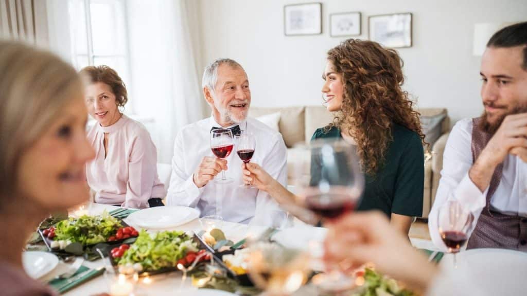 A smiling older man in a bowtie toasts a young woman at a dinner table with other people.