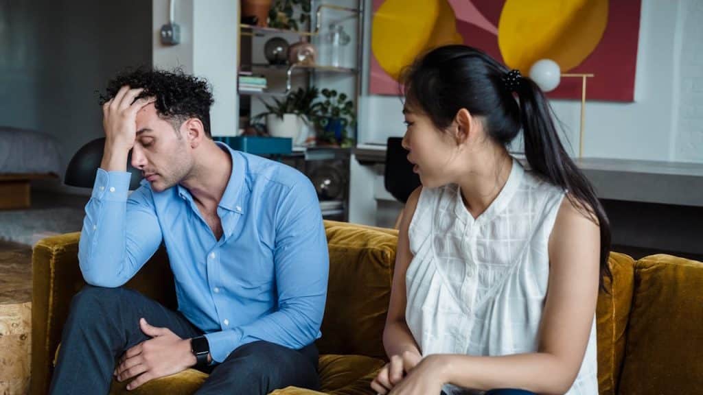 A couple is seated on a couch during a serious argument, with the man looking stressed with his hand on his head.