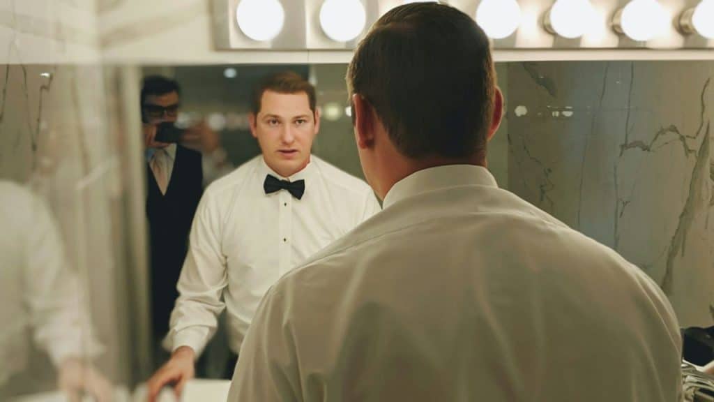 A man in a bow tie looks at his reflection in a brightly lit bathroom mirror.