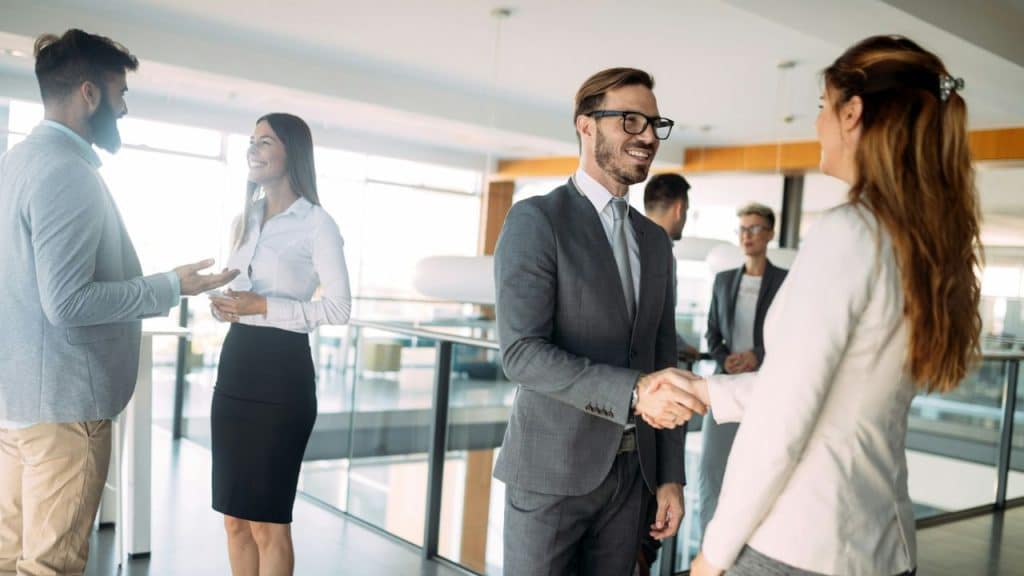A group of business professionals is seen networking in a modern office, with one man and woman shaking hands in the foreground.