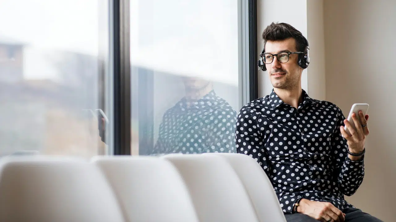 A man wearing glasses and headphones is looking out a window while holding a phone.