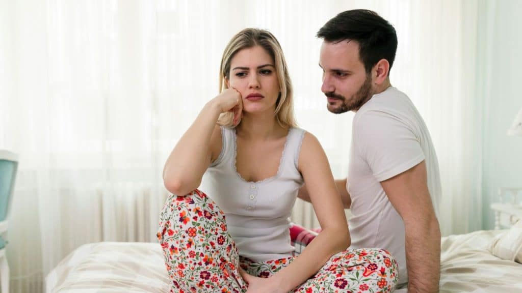 Upset woman in floral pants sitting on a bed while a man looks at her with concern.
