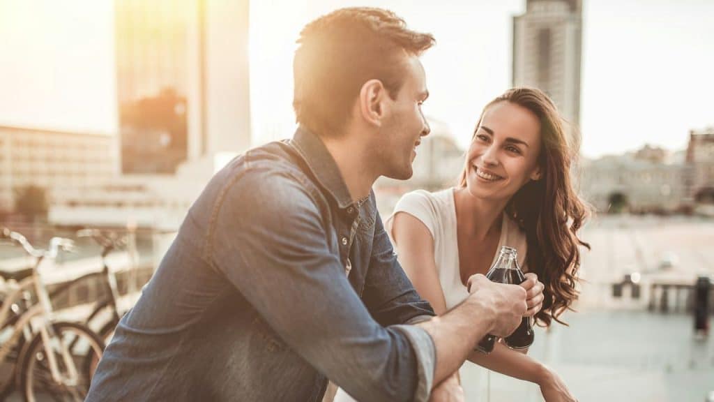 A smiling man and woman are talking and holding a bottled drink on a sunny city balcony.