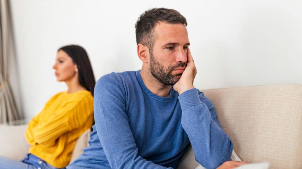 A distressed man in a blue sweater sits in the foreground, with his chin resting on his hand, while a woman sits turned away from him in the background.