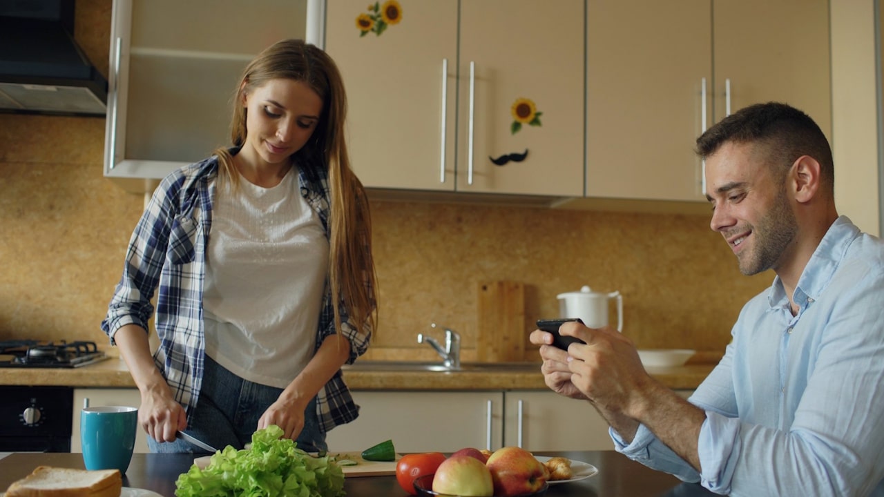 A woman is cutting vegetables in the kitchen while her husband is playing game on mobile phone.