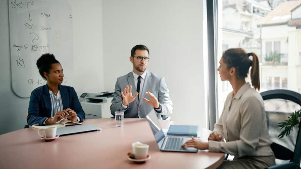 Three colleagues discussing at a round table, man in center gesturing with his hands.