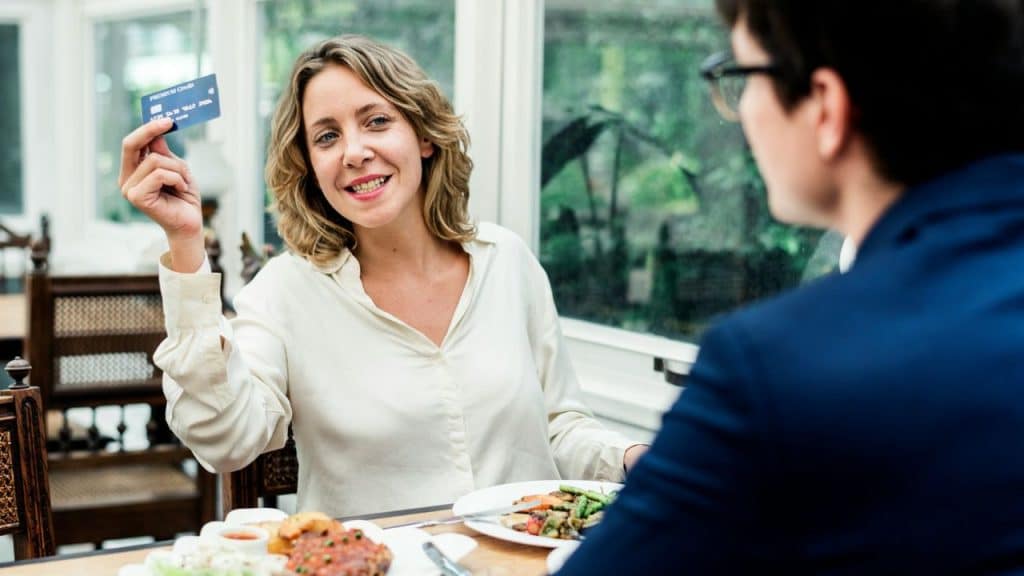 A smiling woman holds up a credit card across the table from a man in a suit.