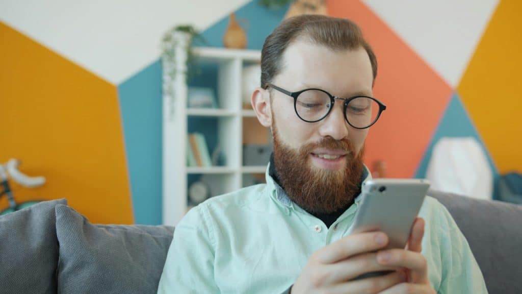 A man with a full red beard and round black glasses smiles while looking down at his smartphone, sitting on a couch.