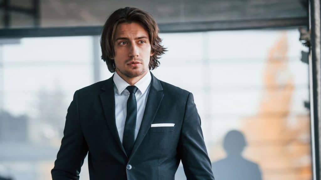 Young man with long, dark hair wearing a black suit and tie indoors, looking off to the side.
