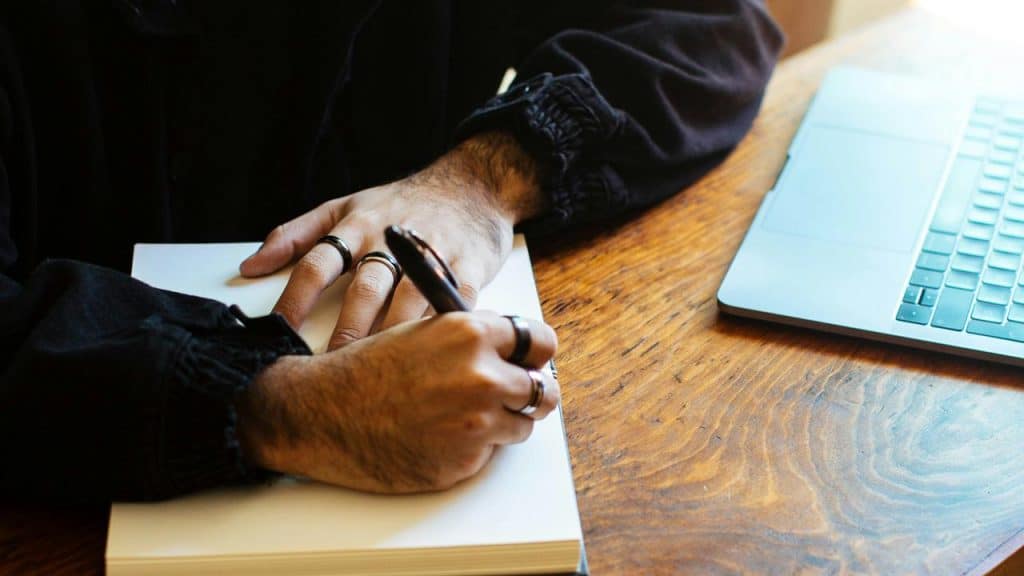 A person writing in a notebook beside a laptop on a wooden desk.