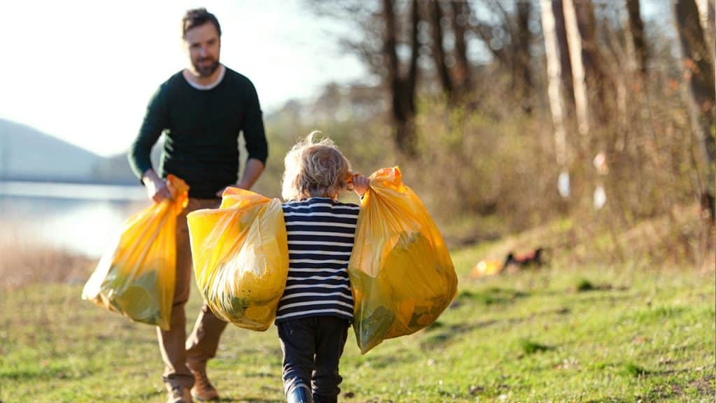 A child and an adult carrying large bags of trash across a grassy area outdoors.