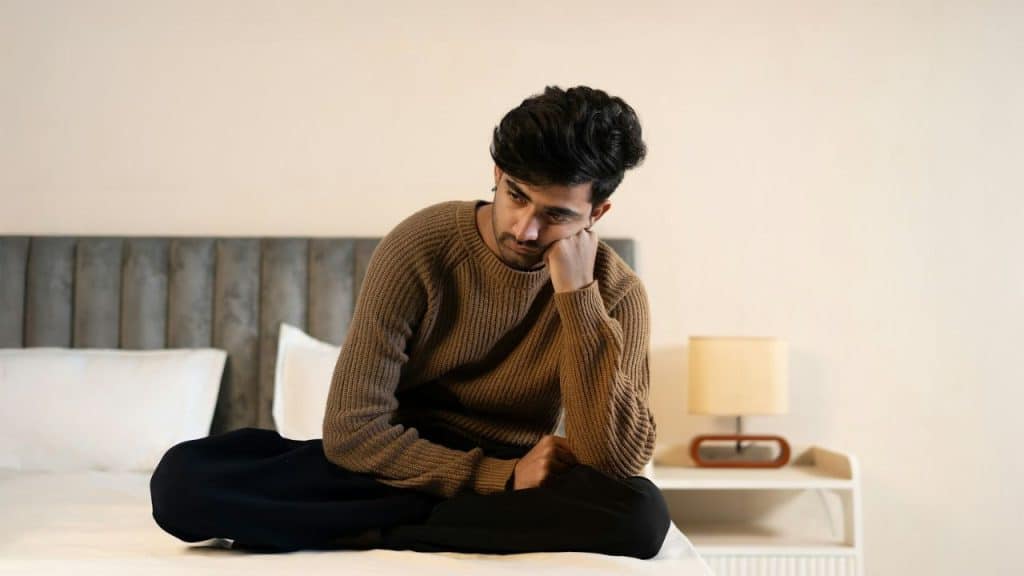 Young man in a brown sweater sitting on a bed, looking down with a hand on his cheek.