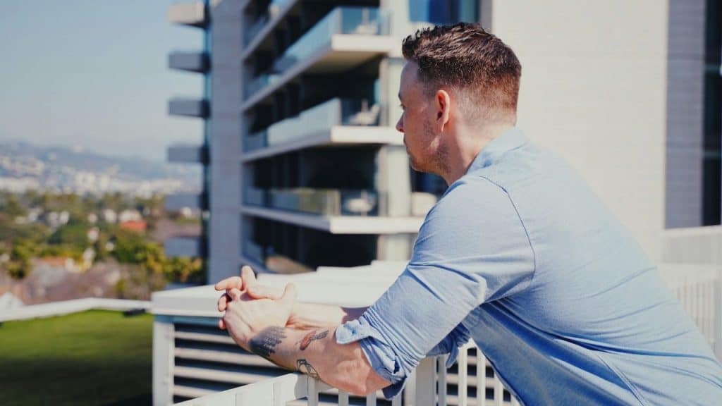 Man with tattoos on his forearm looking out from a high-rise balcony on a sunny day.