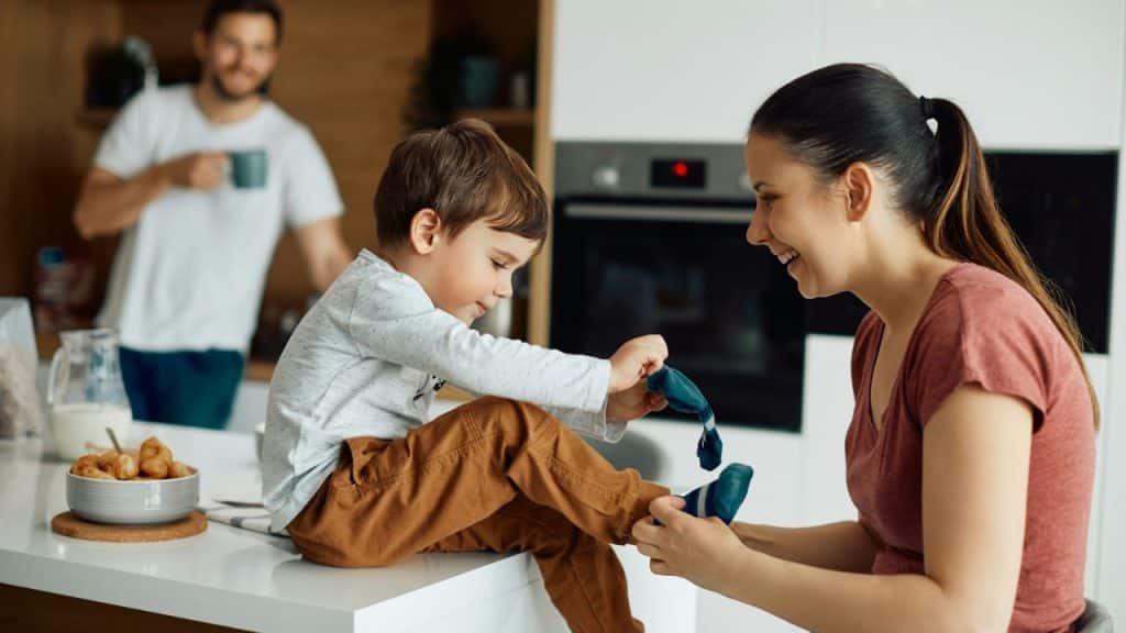 Woman smiling while helping a young boy put on socks in a kitchen, with a man in the background.