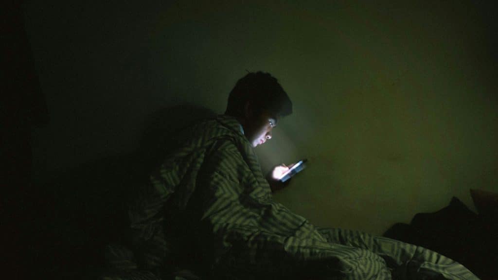 A person is sitting in a dark room, covered with a blanket, looking at a glowing smartphone held in their hands.