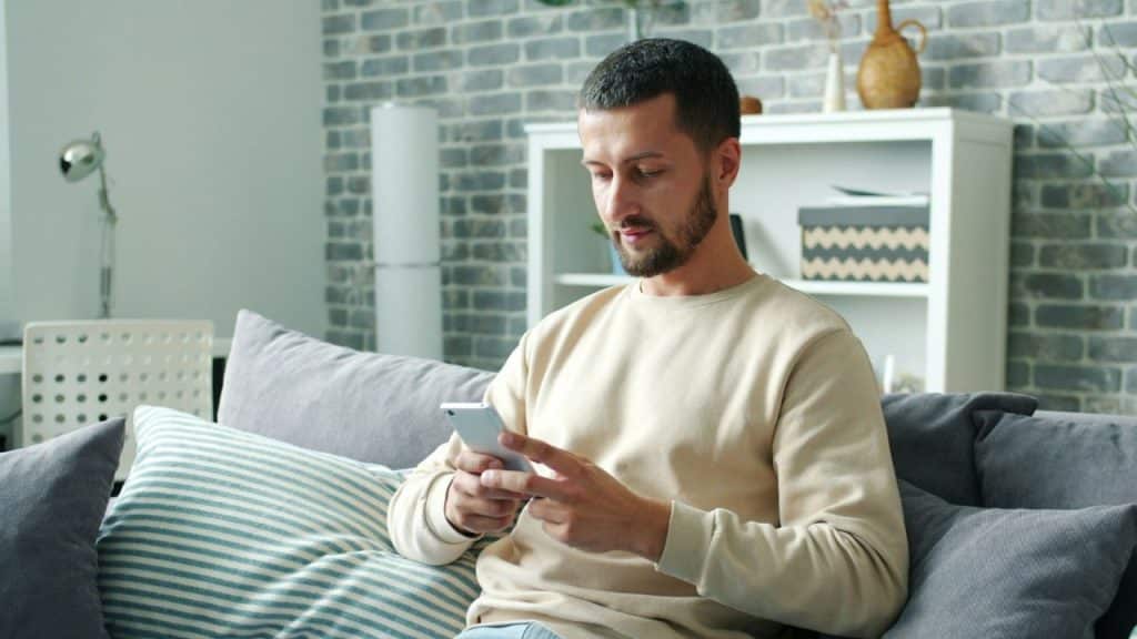 Man in a beige sweater sitting on a couch and looking at his smartphone.