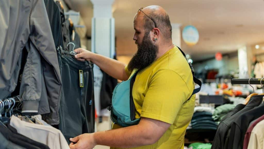 Bald, bearded man in a yellow shirt examining clothes on a rack in a store.