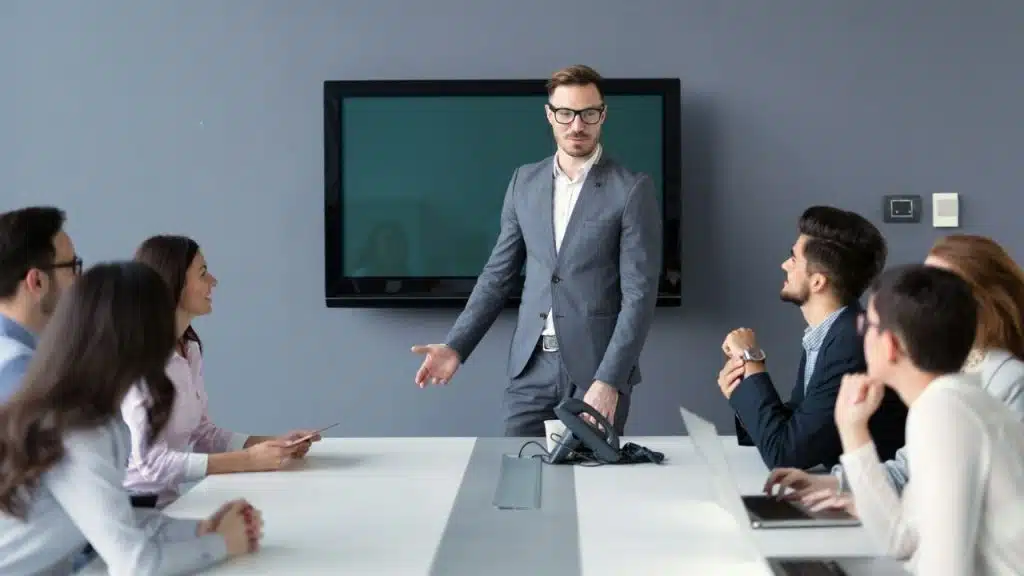 A bespectacled man in a suit addresses colleagues seated around a conference table.