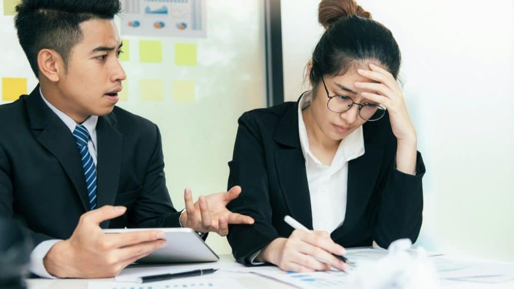 Two professionals at a desk; a man speaking and a woman resting her head on her hand.