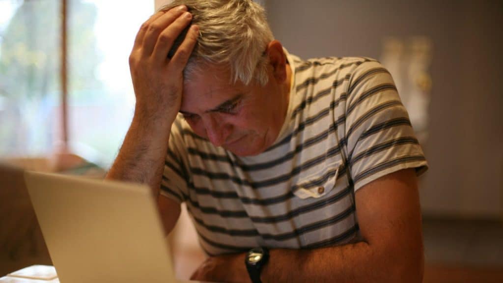 Older man looking distressed over a laptop with his hand on his head.