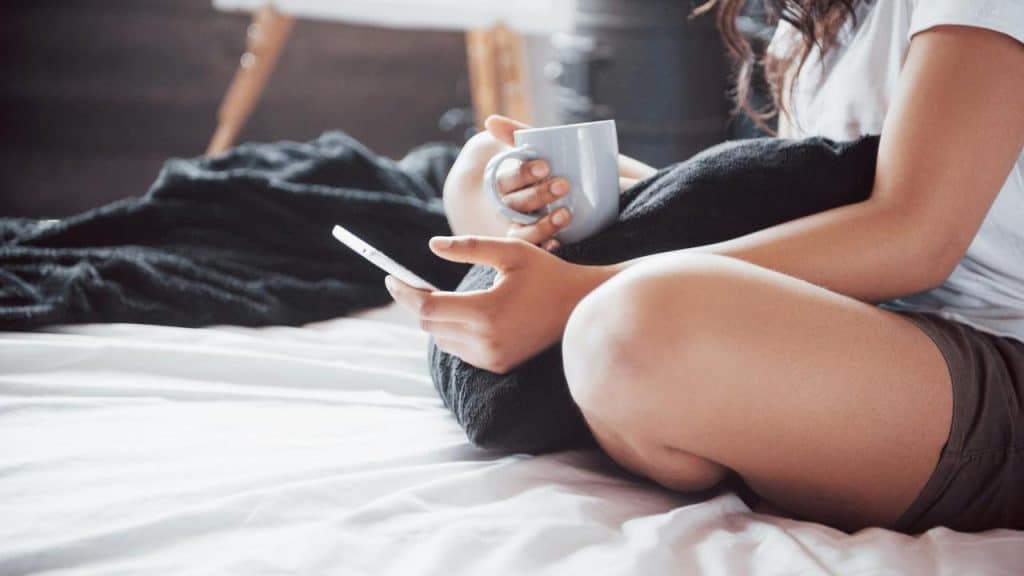 A woman sitting on a bed holding a mug and a phone.