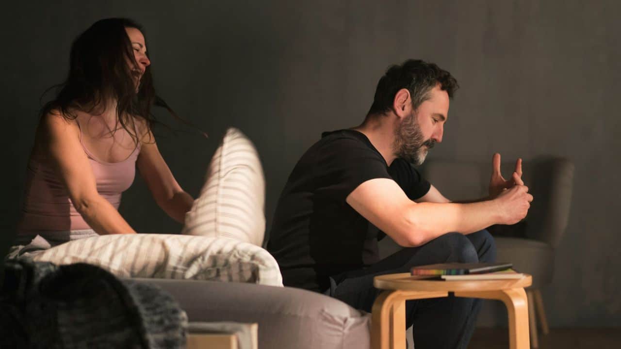 Woman in background yelling while hitting a man with a pillow on a couch.